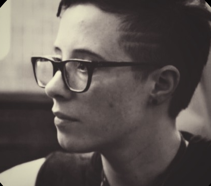 Portrait of Alex Zaremba in a black-and-white photograph, wearing thick, dark rectangular glasses and a dark top